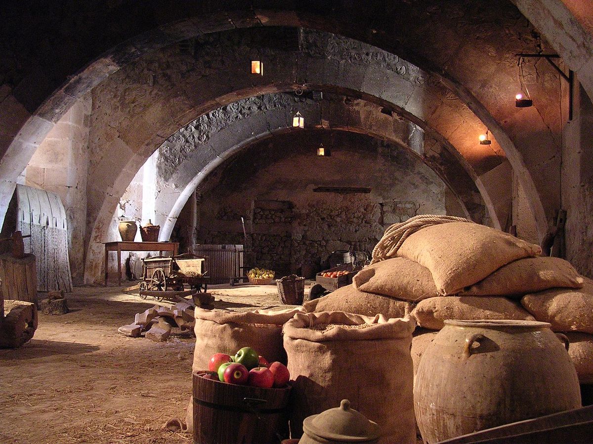 Bodega abovedada de piedra con sacos, barriles y una caja de manzanas.