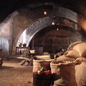 Bodega abovedada de piedra con sacos, barriles y una caja de manzanas.