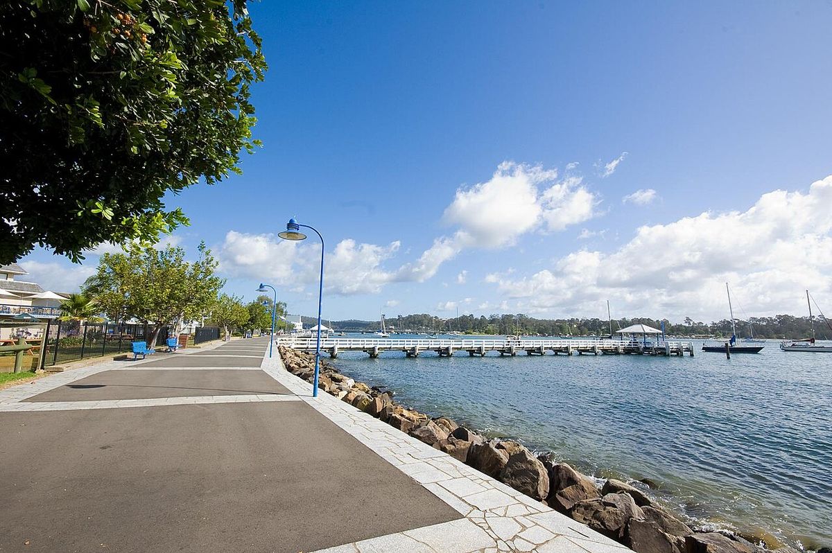 Paseo junto al mar con árboles a un costado y embarcaciones a lo lejos.