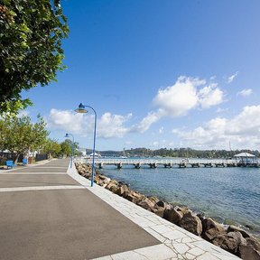 Paseo junto al mar con árboles a un costado y embarcaciones a lo lejos.